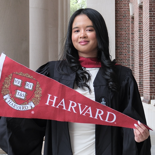 SRJC alumna Kanto Raveloson smiles at the camera during her graduation from Harvard University. She wears a black graduation gown and holds a red Harvard pennant. 