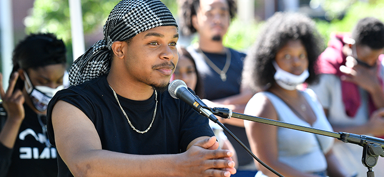 A young African American man gives a speech during an outdoor event at Santa Rosa Junior College. Behind him, blurred, are several young African American male and female students.