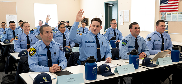 Male and female students in the Basic Police Academy are sitting at their desks, ready to respond to the instructor. They are wearing light blue uniforms with shoulder patches that indicate their respective police departments. 