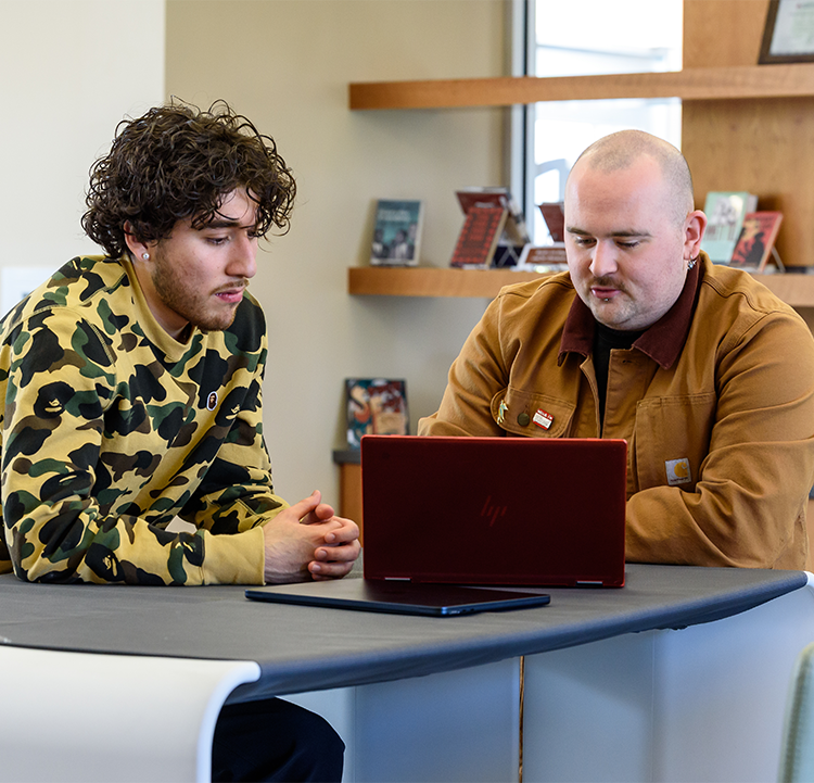 Two male students are looking at a laptop in the library at Santa Rosa Junior College – Petaluma Campus. One of them has curly hair and wears a camouflage sweatshirt. The second is bald and wears a beige light jacket. 