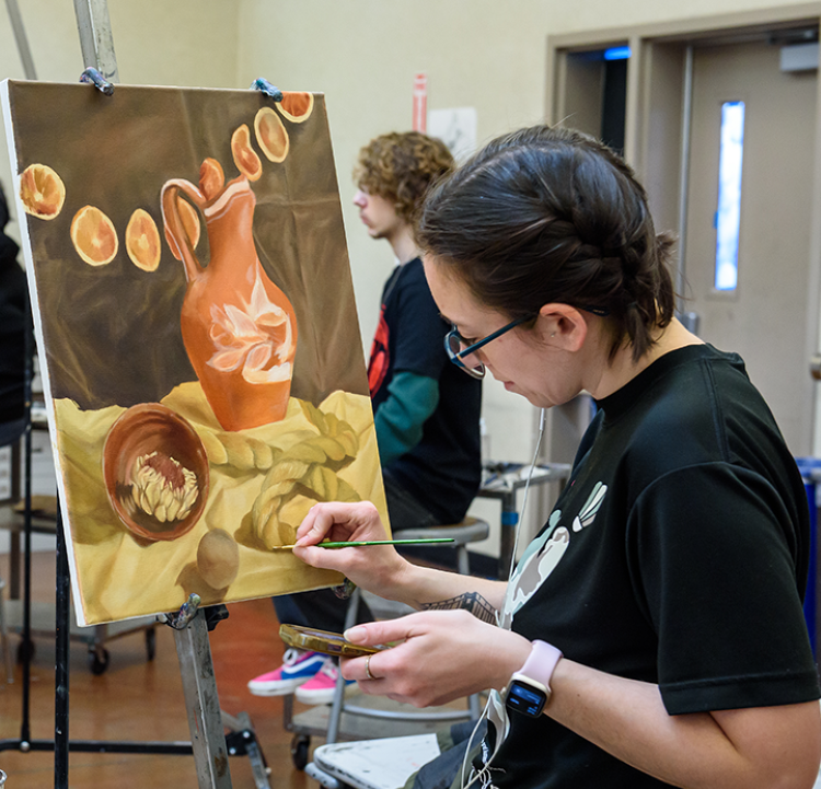 A woman with short dark hair wearing a black T-shirt paints a ceramic carafe and a bowl on canvas during a painting class at Santa Rosa Junior College. 