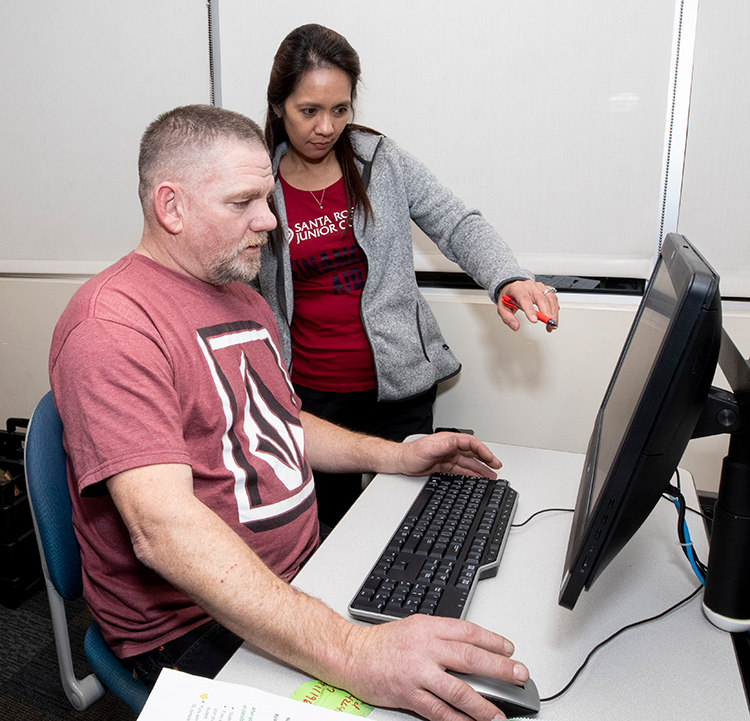 A female employee wearing a red SRJC Financial Aid t-shirt assists a male returning student seated at a computer. 