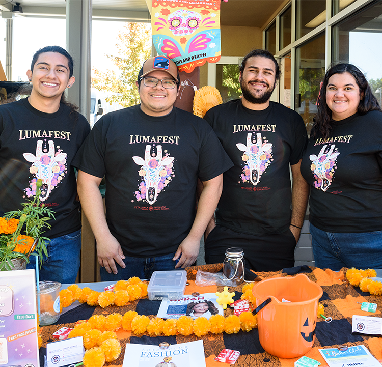Four students (three male and a female) tabling during the LumaFest event at SRJC Petaluma are smiling at the camera. They wear black T-shirts with a giraffe skeleton head and the inscription LumaFest.