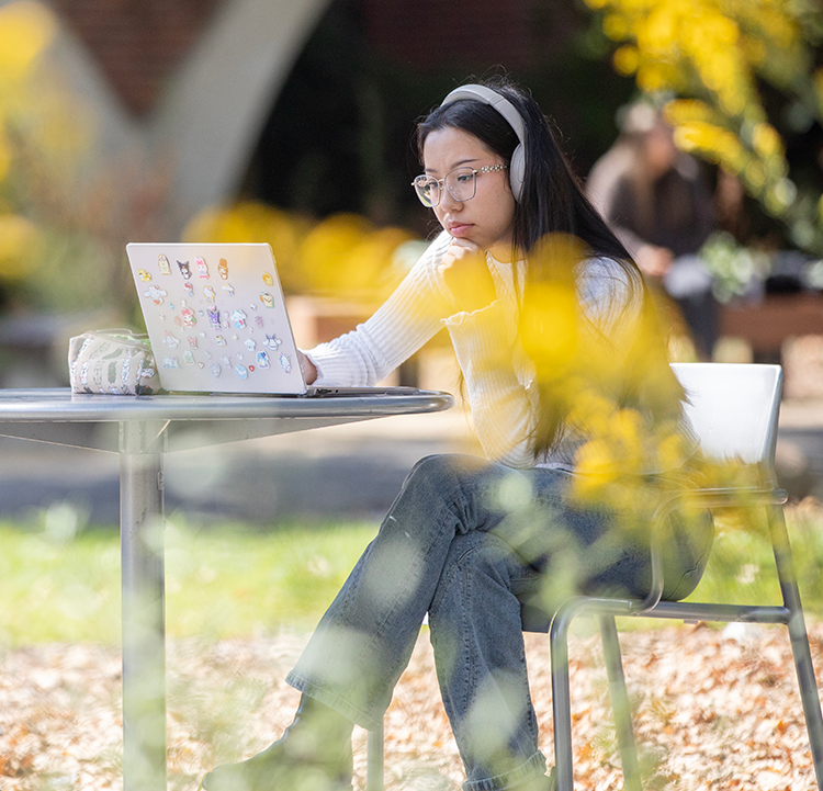 A young woman with long black hair wearing big white headphones is watching a laptop. She is seated at an outside table. Her laptop is covered in cute animal stickers.  