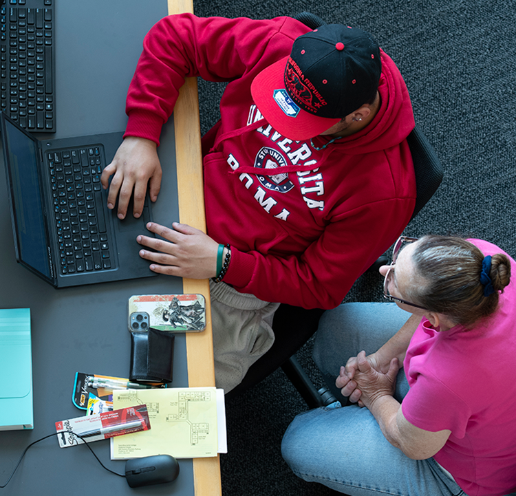 A seated male student is working on a laptop assisted by a seated female staff member in the library of Santa Rosa Junior College.