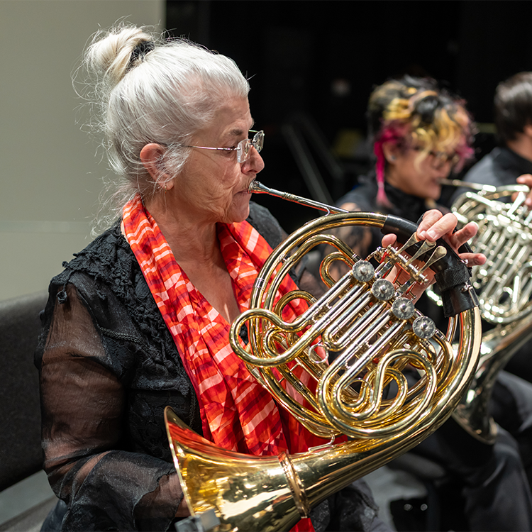 A lifelong learner female student wearing evening black attire and a red scarf is playing the horn during a performance with the SRJC Symphonic Orchestra.