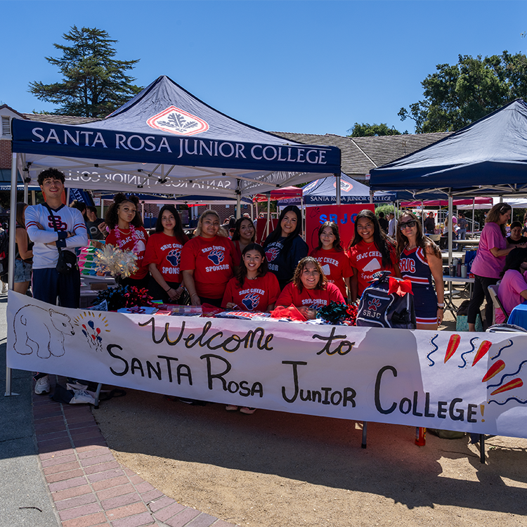 A group photo of the SRJC Cheer team in red t-shirts, lined up behind a table in the quad at Santa Rosa Junior College. The tablecloth reads “Welcome to Santa Rosa Junior College. Behind the group is a blue canopy displaying the college’s name and the acorn leaf logo. 