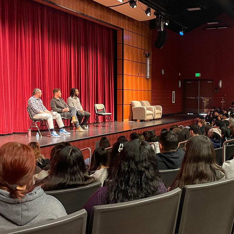 An image of a conference held at the Ellis Auditorium at Santa Rosa Junior College. Three men are seated on a stage, in front of a red velvet curtain. They are listening to someone from the audience who interacts with them. 