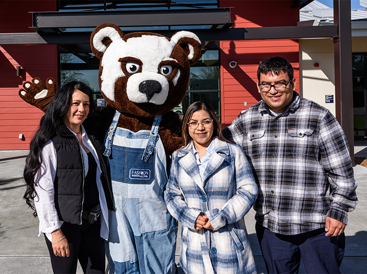 Three students, two female and a male, pose with Rosco the bear cub, the Santa Rosa Junior College mascot, in front of SRJC Roseland center. 