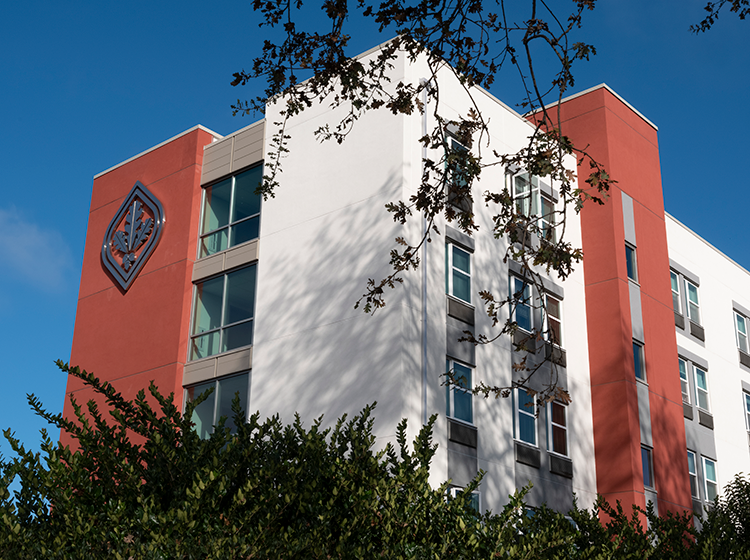 A photo of the Polly Hall Student Housing on the Santa Rosa campus of SRJC against the blue sky. The building is brick red and white, has the oak leaf logo on the upper left corner and is framed by greenery and oak tree leaves. 