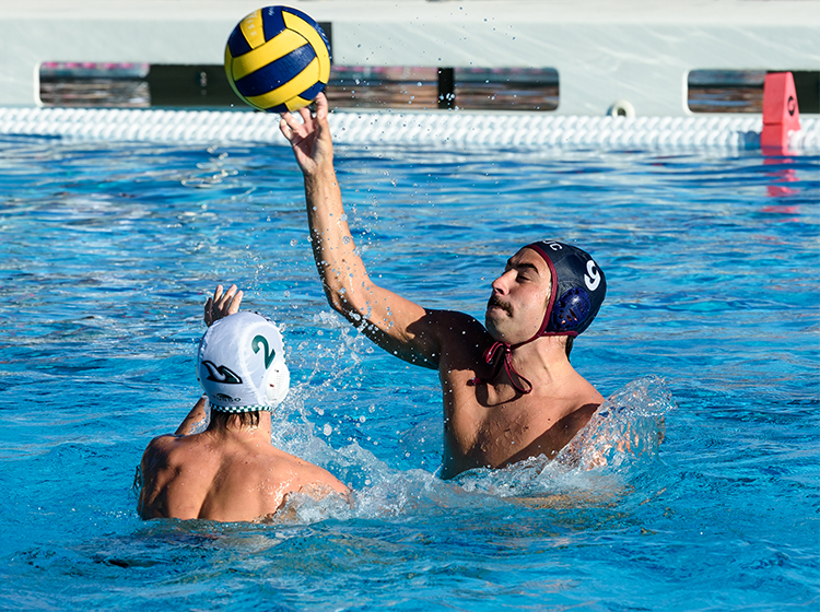 Two athletic young men are playing water polo in the Olympic pool at Santa Rosa Junior College. One is wearing a navy-blue SRJC water polo cap with red accents and the number 9, and is holding a yellow-and-black ball above his head, ready to throw it over the opponent’s reach.  