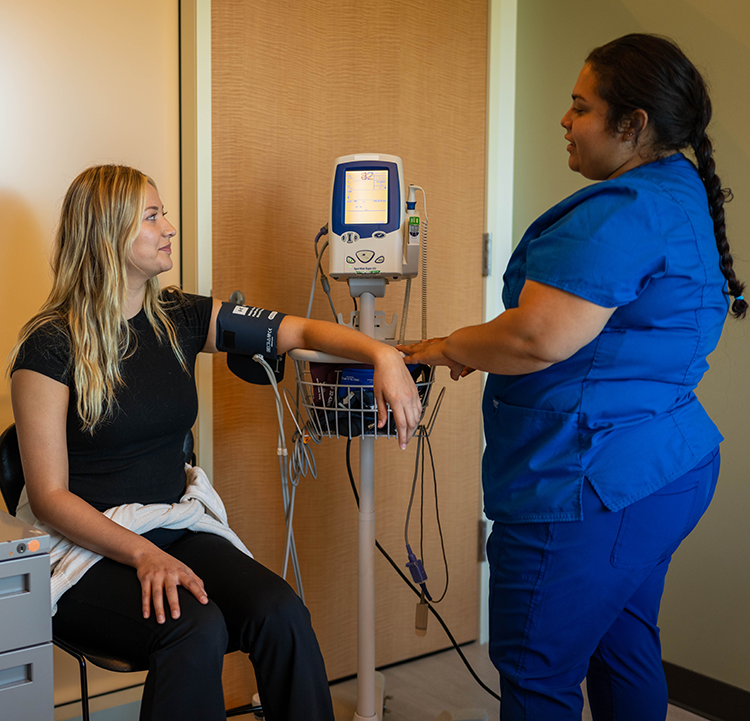 A female nurse wearing blue scrubs is taking the blood pressure of a female student in a medical cabinet at Santa Rosa Junior College