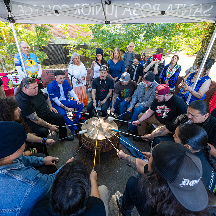 Indigenous male and female performers sing in a drum circle under a canopy tent at Santa Rosa Junior College. 