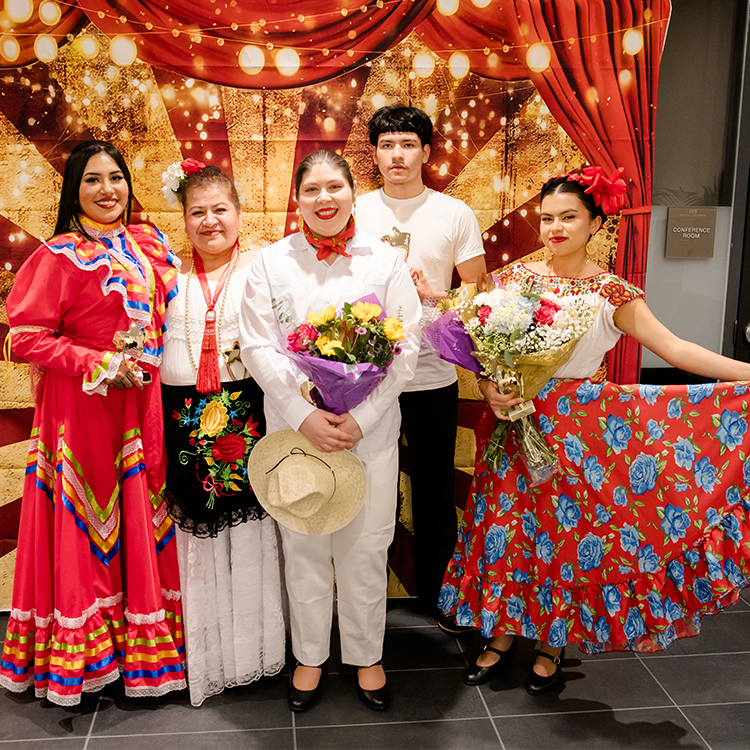 A group of male and female Latine performers smiles at the camera on the stage of La Carpa event at Santa Rosa Junior College. They are wearing multicolored attire that represents several traditional dances and regions of Mexico. 