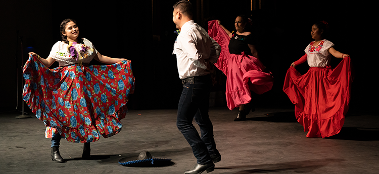 A man and three women perform a Ballet Folclórico dance on the stage at Santa Rosa Junior College. The women are wearing traditional Latinx attire with colorful long skirts and decorated blouses and the man wears black pants and a white shirt.