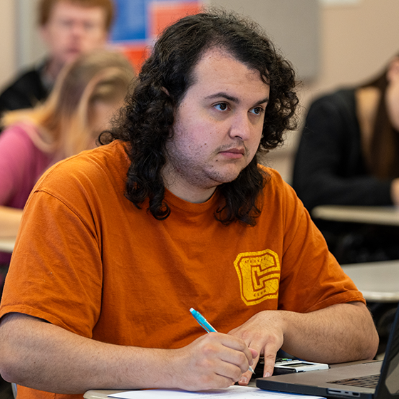 A young male with dark curly hair wearing an orange t-shirt takes notes in a Mathematics classroom at Santa Rosa Junior College. 