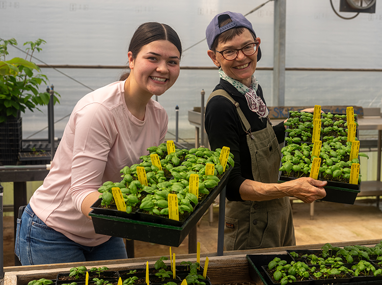 Two female students show the camera trays filled with veggie starters. The female on the left wears a pink shirt and jeans, and the one on the right wears brown overalls over a black shirt. 