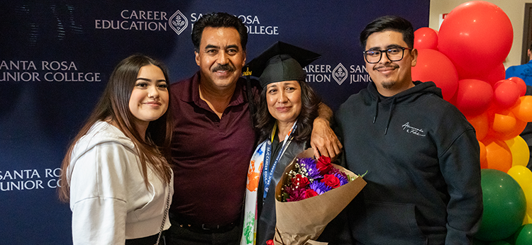 A Latinx family with two young adult children (one male and one female) celebrates the achievements of the mother and wife, during the Celebrate Career Education event at Santa Rosa Junior College. The four people pose indoors against a navy-blue college photo background. 