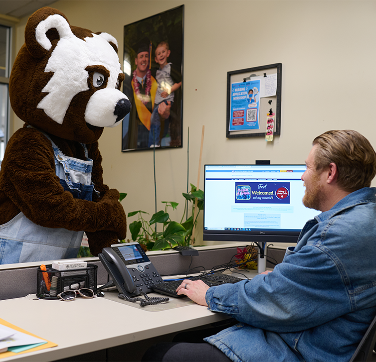 Rosco the Bear Cub, the SRJC mascot, stands at a desk in the Welcome and Connect Center, where a male employee seated at a computer helps Rosco register. 