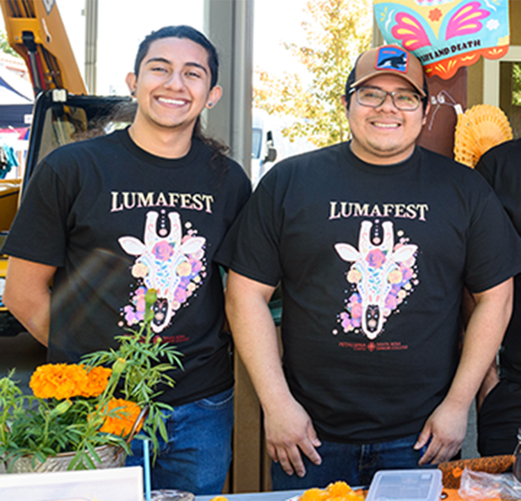 Students tabling during the LumaFest event at SRJC Petaluma are smiling at the camera. They wear black T-shirts with a giraffe skeleton head and the inscription LumaFest.