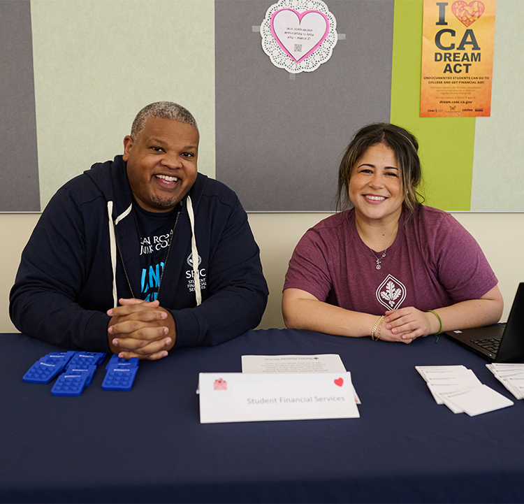 Two Santa Rosa Junior College Financial Aid Specialists, a black male and a Latina female, are smiling at the camera while sitting at a table during a Financial Aid Fair. 