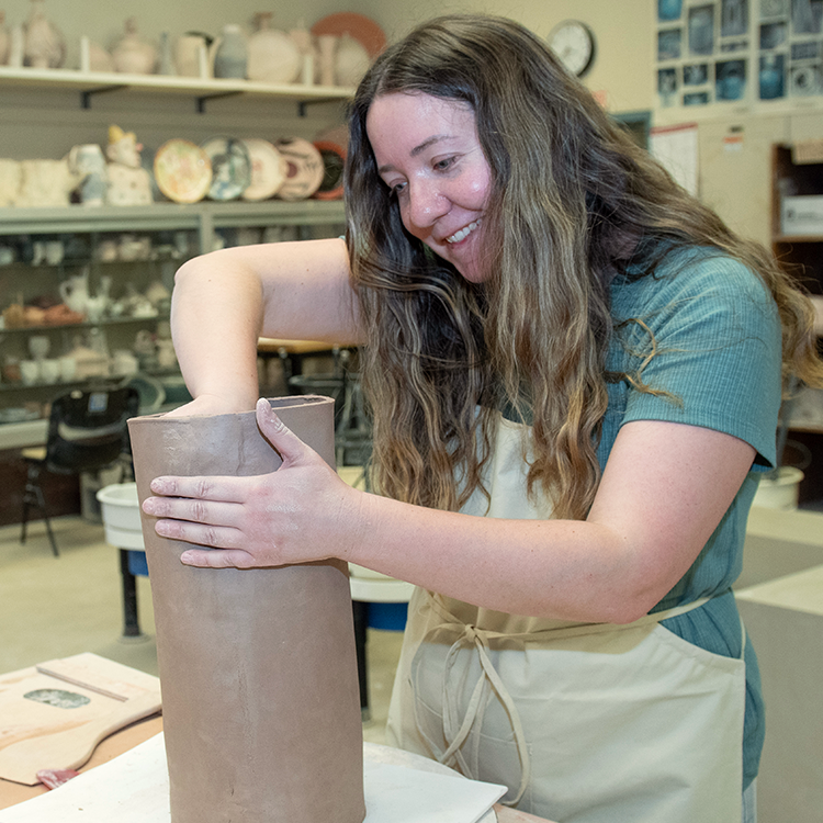 A female with long blond hair wearing a white apron over her clothes is working on a round clay sculpture in the ceramics lab at Santa Rosa Junior College. Around her are shelves filled with pottery pieces. 
