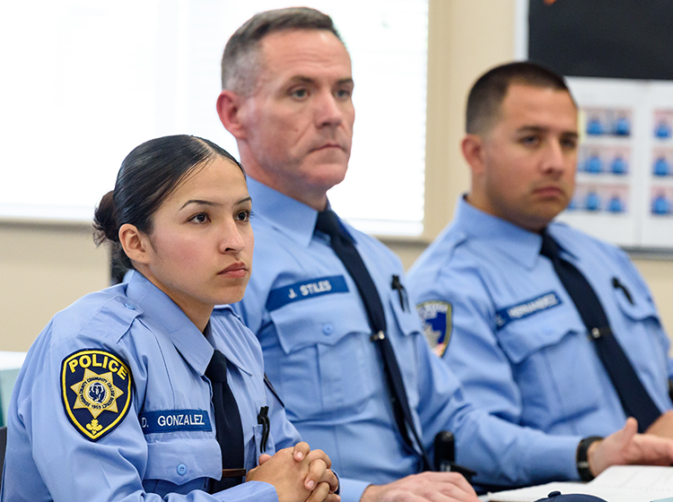 A photo of three police academy students – a female and two males, seated in class and paying attention to a speaker (not shown). They wear light blue police shirts and navy blue ties, and name tags embroidered on their shirts. 
