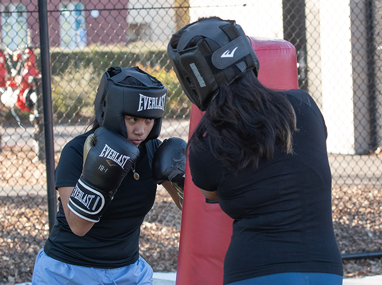 Two female students wearing boxing gloves and head protection gear are punching a boxing bag on the Athletics field at Santa Rosa Junior College.