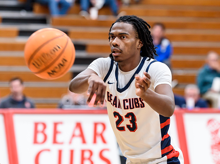 A young black athlete with medium-length dreads passes a basketball during a game at Santa Rosa Junior College. He is wearing a white shirt inscribed with “Bear Cubs 23”. Behind him, multiple people in the stands watch the game. 