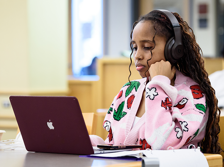 A Black young female wearing a pink sweater with white and red flowers reads on a laptop in the library at Santa Rosa Junior College. She is also listening to music and wearing big black earphones. 