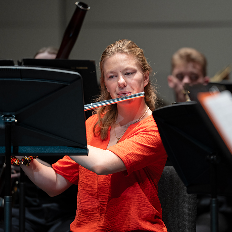 A blonde woman wearing an orange blouse plays the flute during a concert with the Santa Rosa Junior College Orchestra. 